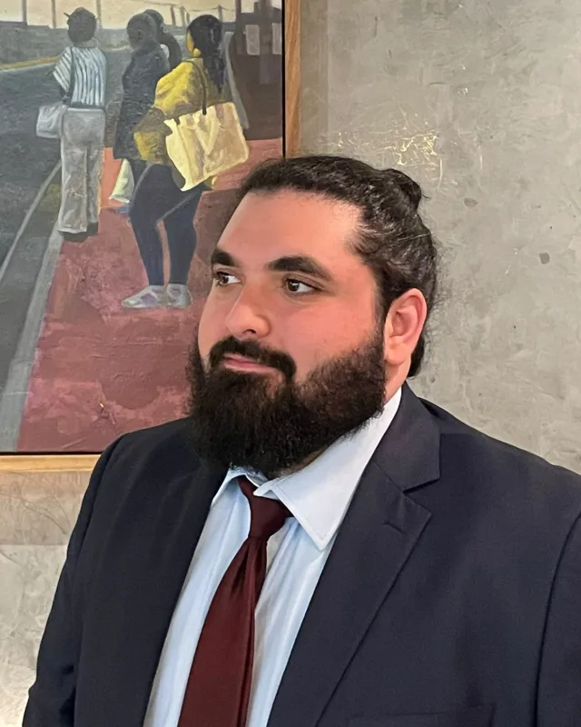 Abdelfattah Abukhmaish in a suit and burgundy tie, looking to the side, beside a framed painting
